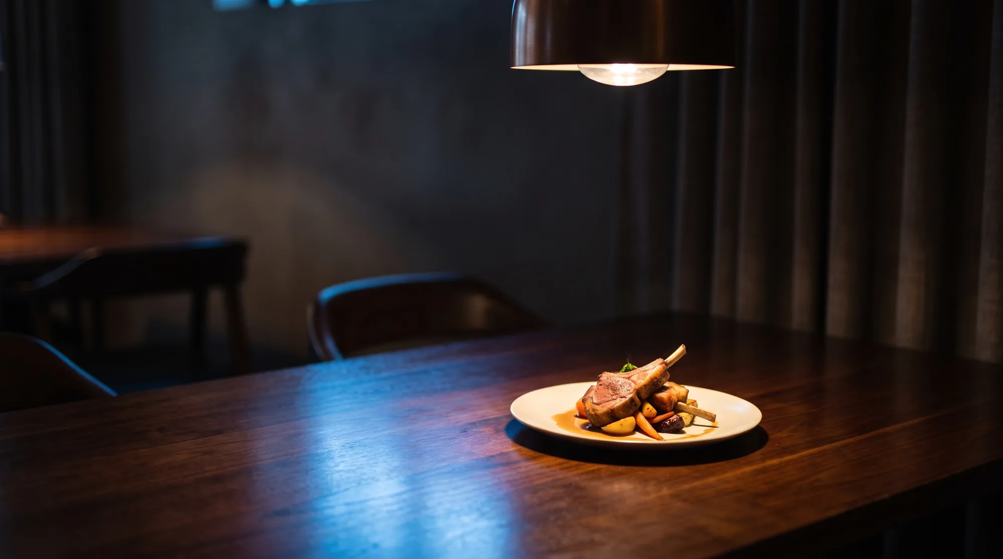 A carefully plated dish sitting alone under a single pendant lamp on a dark restaurant table, the chairs around it empty and the room behind in shadow.
